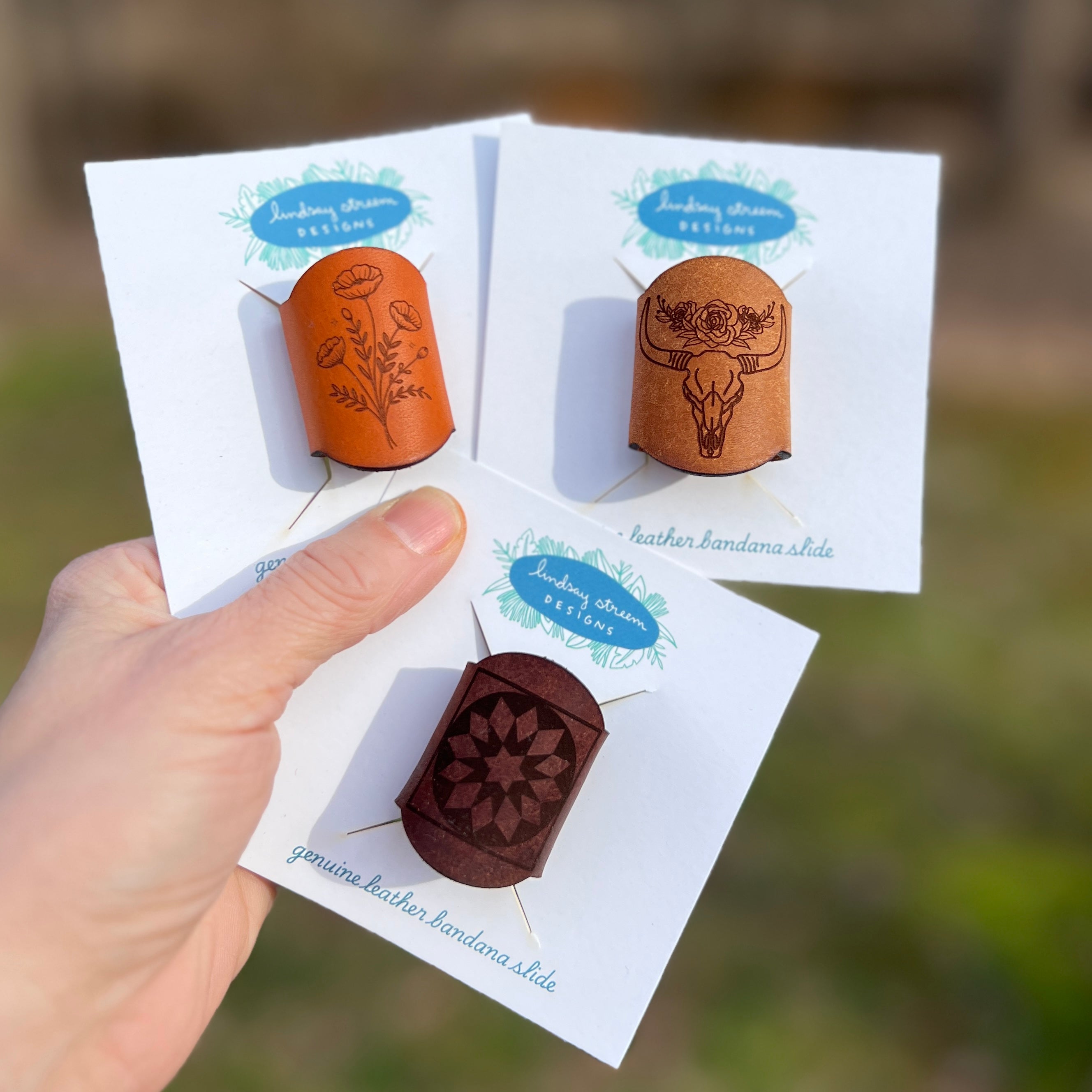 Hand holding three leather bandana slides on cards with a blurred natural background