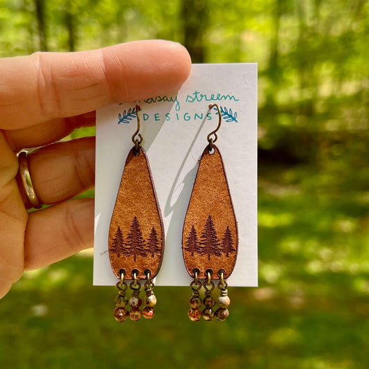 Leather teardrop earrings with tree design held against a blurred green background.