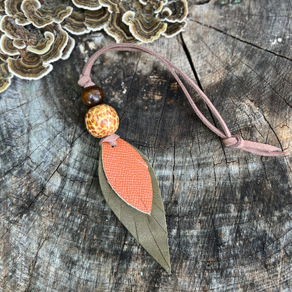 A handmade leather leaf-shaped  keychain or bag charm with a brown cord, patterned orange and beige beads, and a green leather leaf, displayed on a rustic wood background.