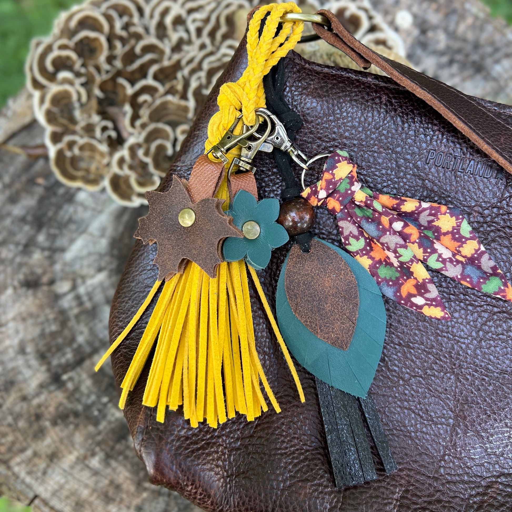 A close-up of a brown textured leather shoulder bag with a decorative keychain or bag charm attached. The charm includes a bright yellow tassel, a brown leather leaf with a green center, a small green flower, and a multicolor floral fabric ribbon.