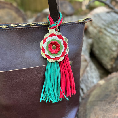 A decorative leather tassel bag charm with a multi-layered flower design in red, green, and gold, hanging from a dark brown leather bag against a blurred outdoor background of rocks.