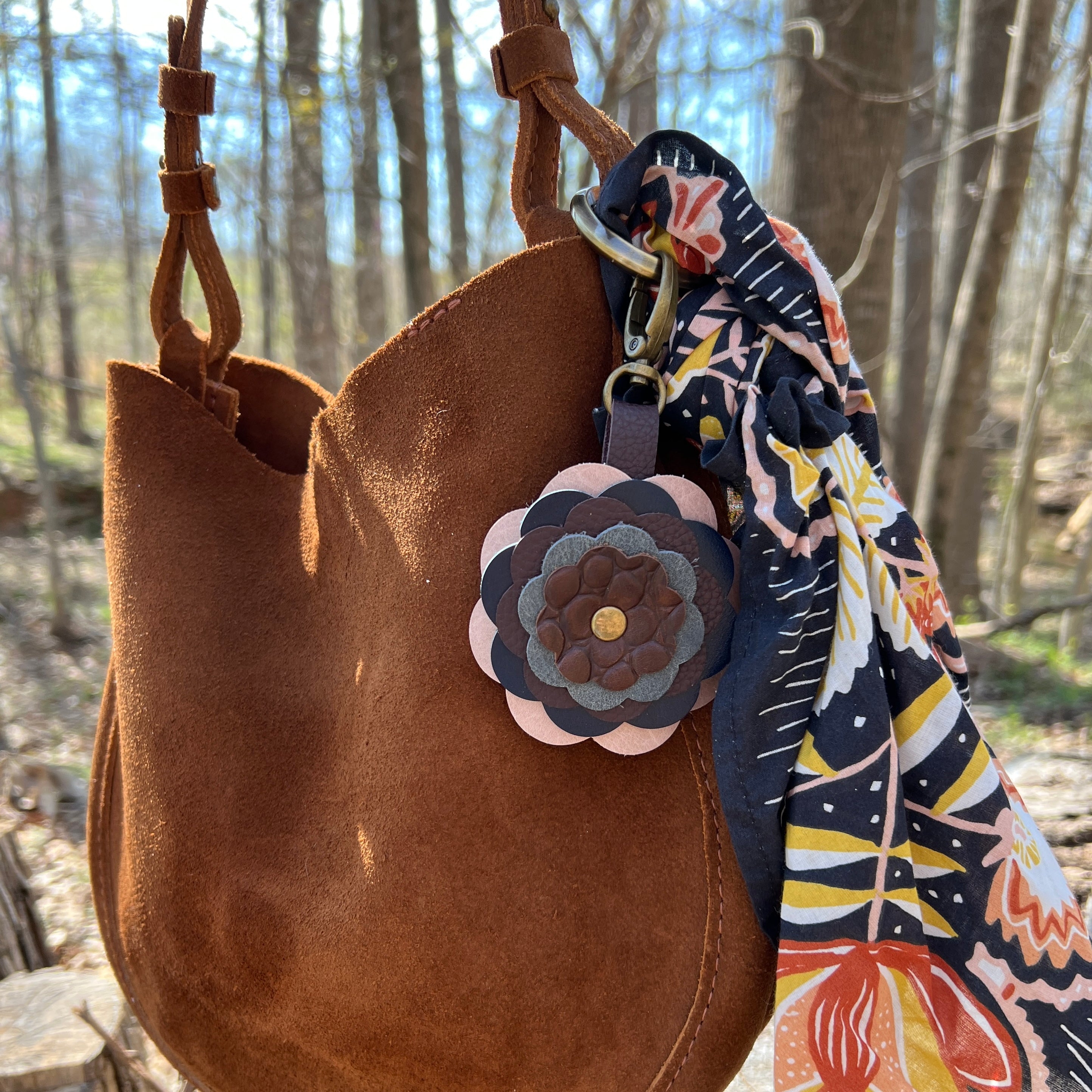 Brown leather bag with floral keychain and scarf in a forest setting