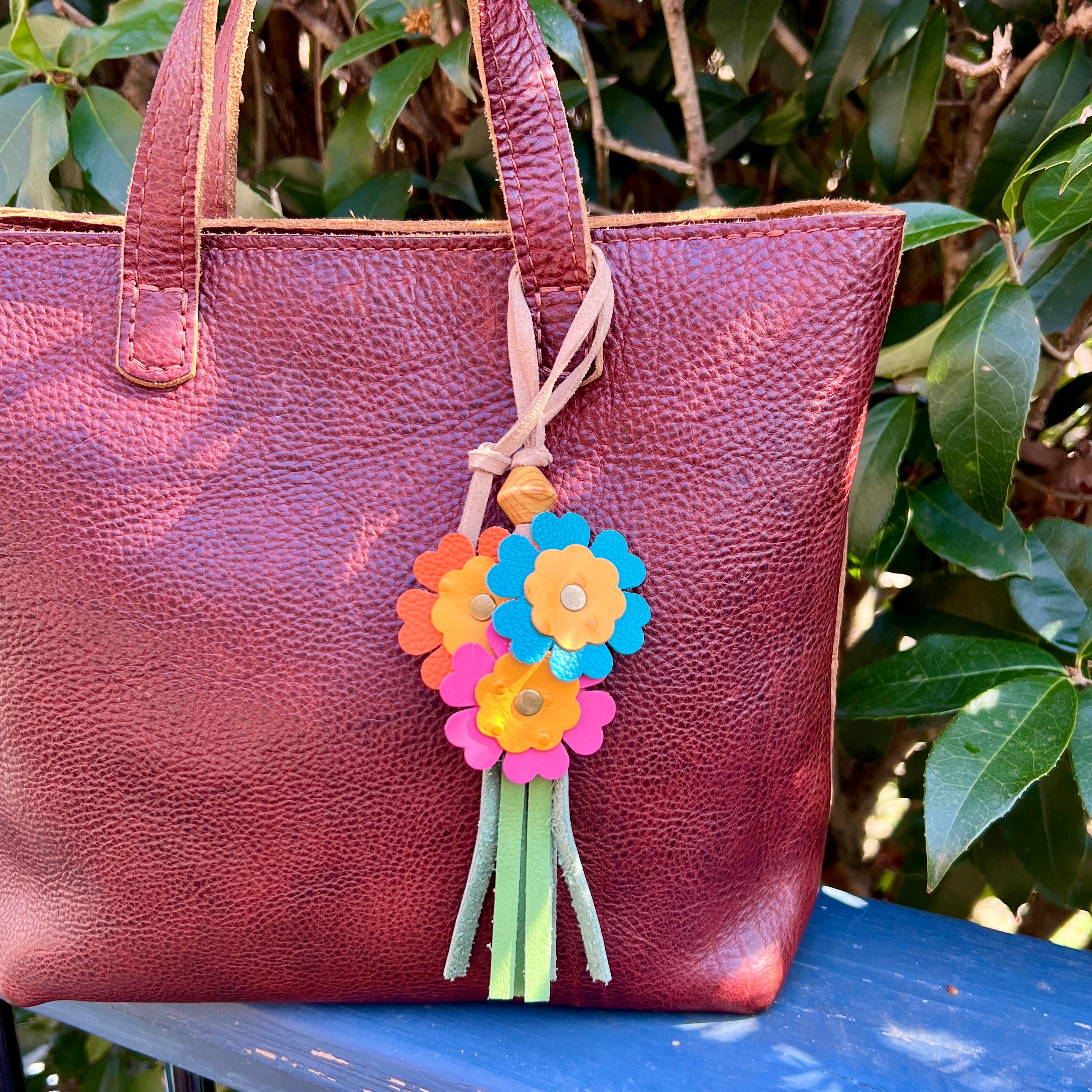 Brown leather tote bag with colorful flower charm against a green leafy background