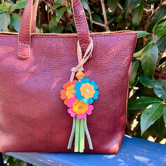 Brown leather tote bag with colorful flower charm against a green leafy background