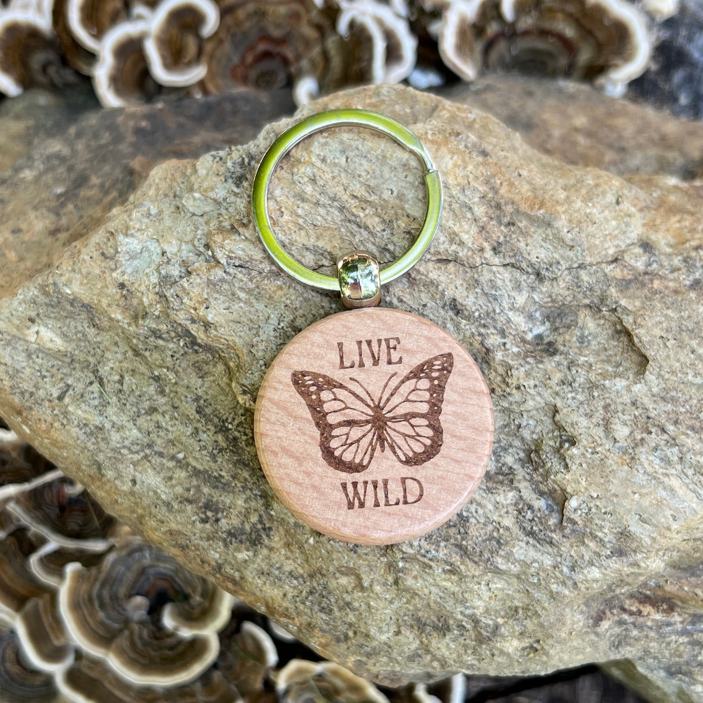 A round wooden keychain tag engraved with a butterfly and the words “LIVE WILD,” attached to a silver split ring and resting on a textured rock with mushrooms in the background.