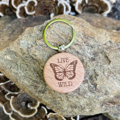 A round wooden keychain tag engraved with a butterfly and the words “LIVE WILD,” attached to a silver split ring and resting on a textured rock with mushrooms in the background.