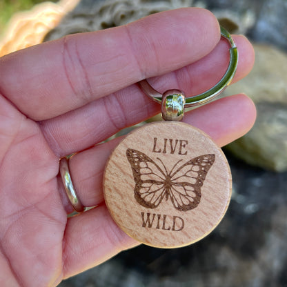 A close-up of a hand holding a round wooden keychain with a butterfly engraving and the words 'LIVE WILD' on a metal key ring, with a blurred natural background.