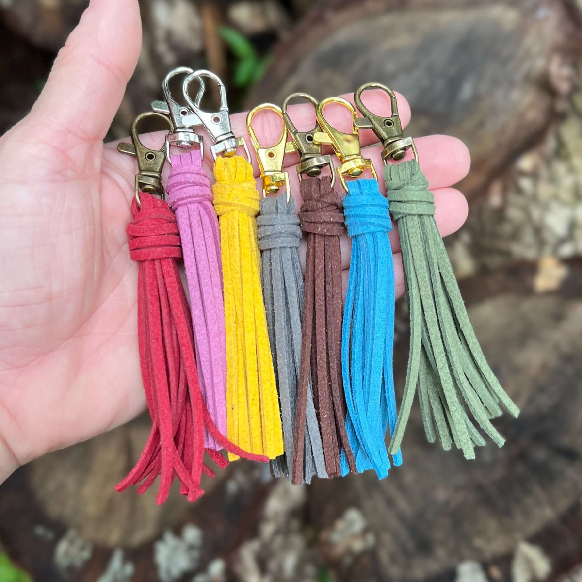 Six suede tassel keychains in red, pink, yellow, gray, brown, and blue, held in a person's hand against a blurred outdoor background of rocks and foliage.