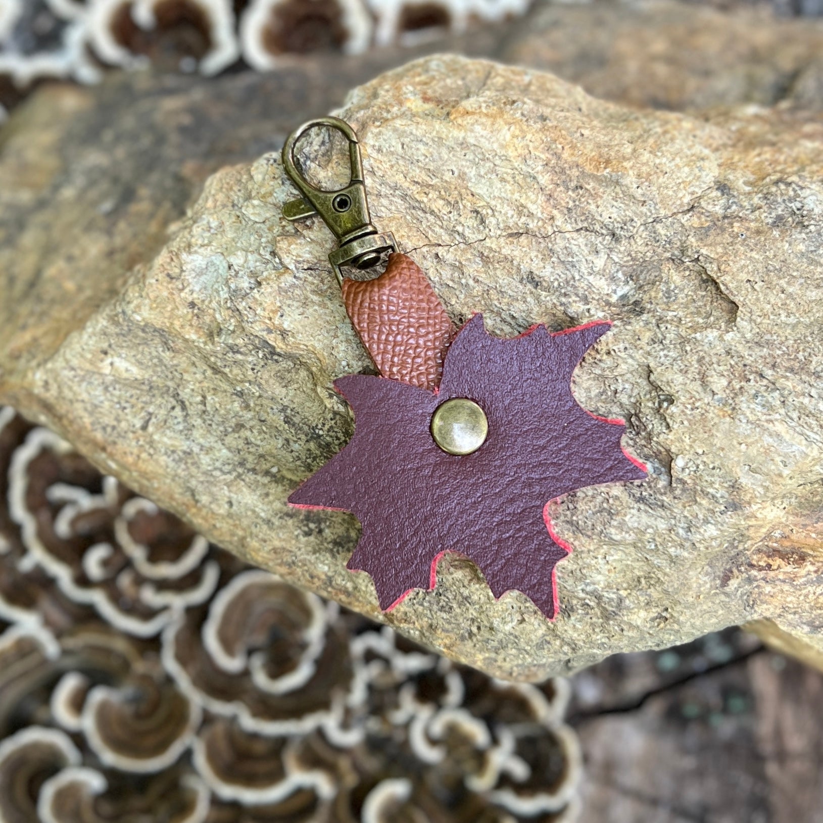 A small handmade leather accessory with a split brown and burgundy leaf-shaped piece, a brass rivet, and an antique bronze lobster clasp, resting on a textured rock.