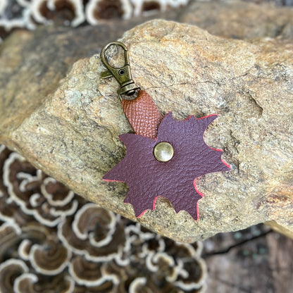 A small handmade leather accessory with a split brown and burgundy leaf-shaped piece, a brass rivet, and an antique bronze lobster clasp, resting on a textured rock.