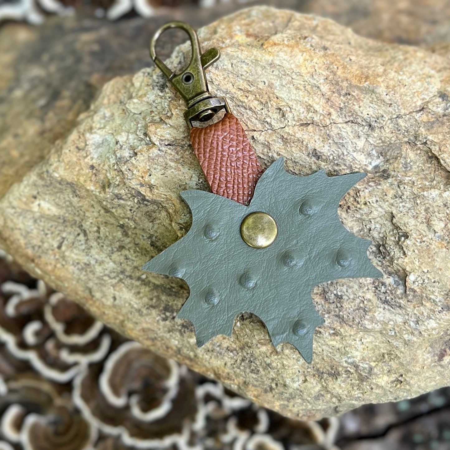 A handmade leather keychain shaped like a leaf, featuring a muted sage green body with a central brass rivet, a reddish-brown textured leather loop, and an antique bronze clasp, displayed on a natural rock.