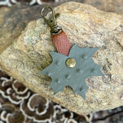A handmade leather keychain shaped like a leaf, featuring a muted sage green body with a central brass rivet, a reddish-brown textured leather loop, and an antique bronze clasp, displayed on a natural rock.