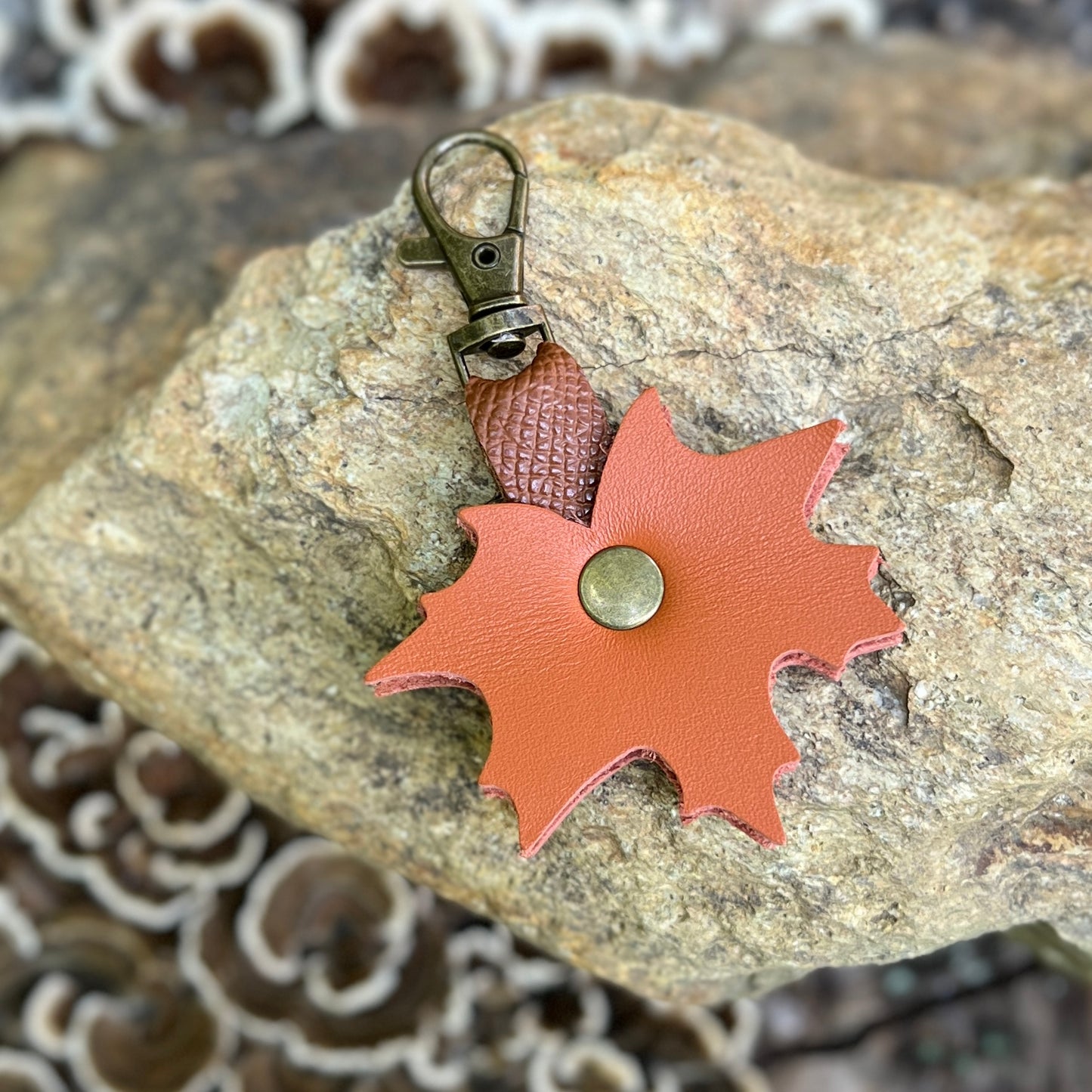 A small orange leather bag charm shaped like a maple leaf, with a brass snap button and an antique bronze lobster clasp, resting on a textured rock.