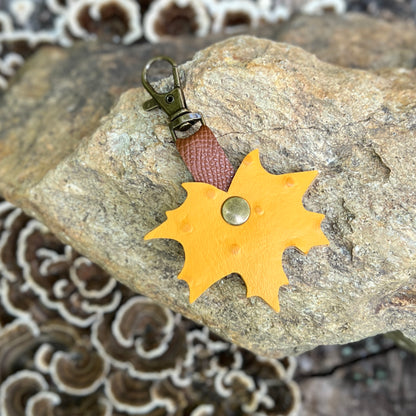 A yellow autumn leaf-shaped leather keychain with a brown textured strap and an antique bronze clasp, resting on a large gray rock with mushrooms in the background.