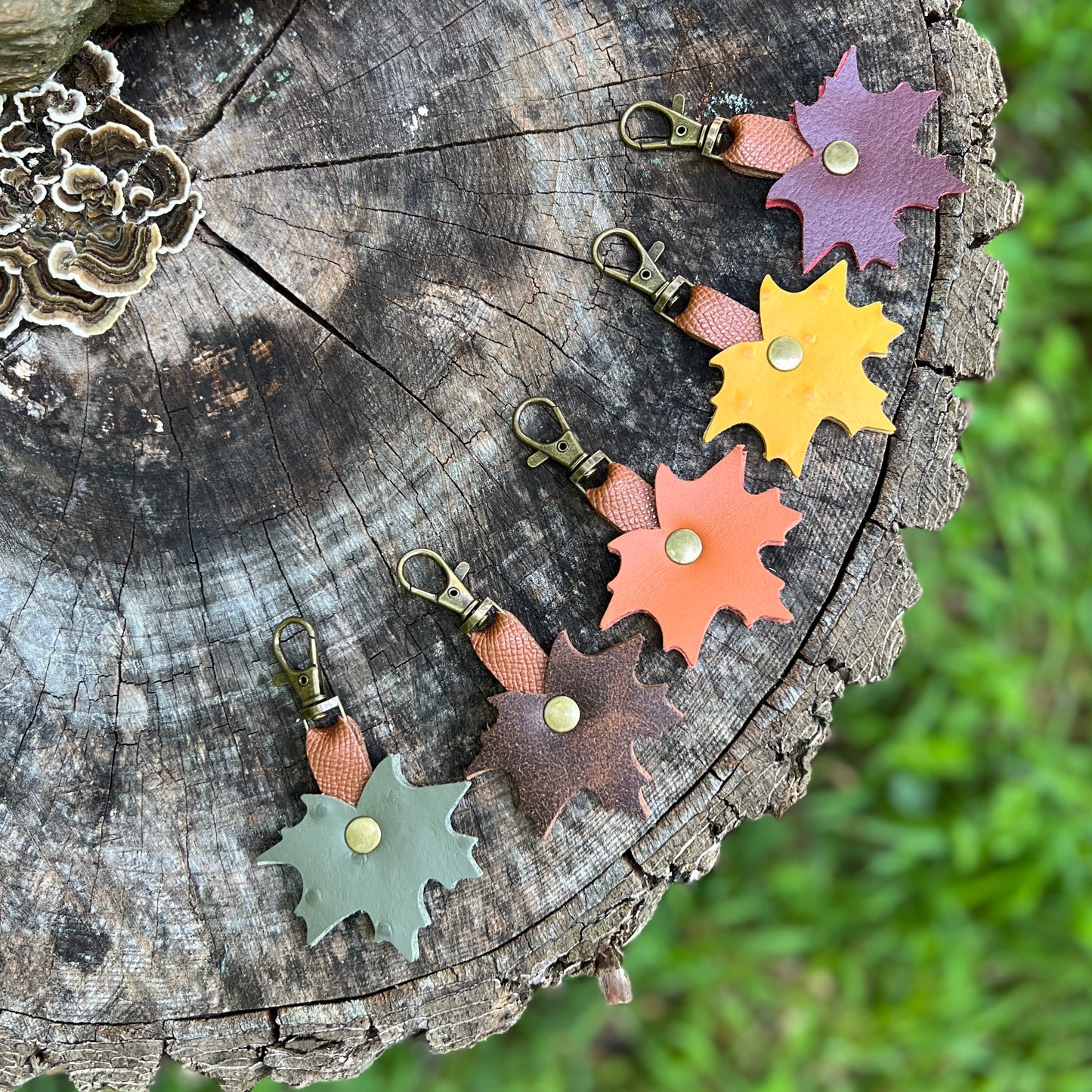 Five colorful leather leaf-shaped keychains with brass snap hooks are arranged on a rustic tree stump outdoors, with blurred green grass in the background.