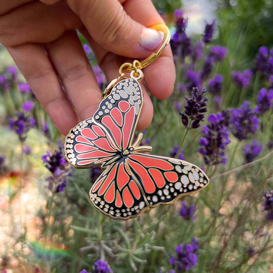 Butterfly-shaped keychain held by a hand with a blurred background of purple flowers