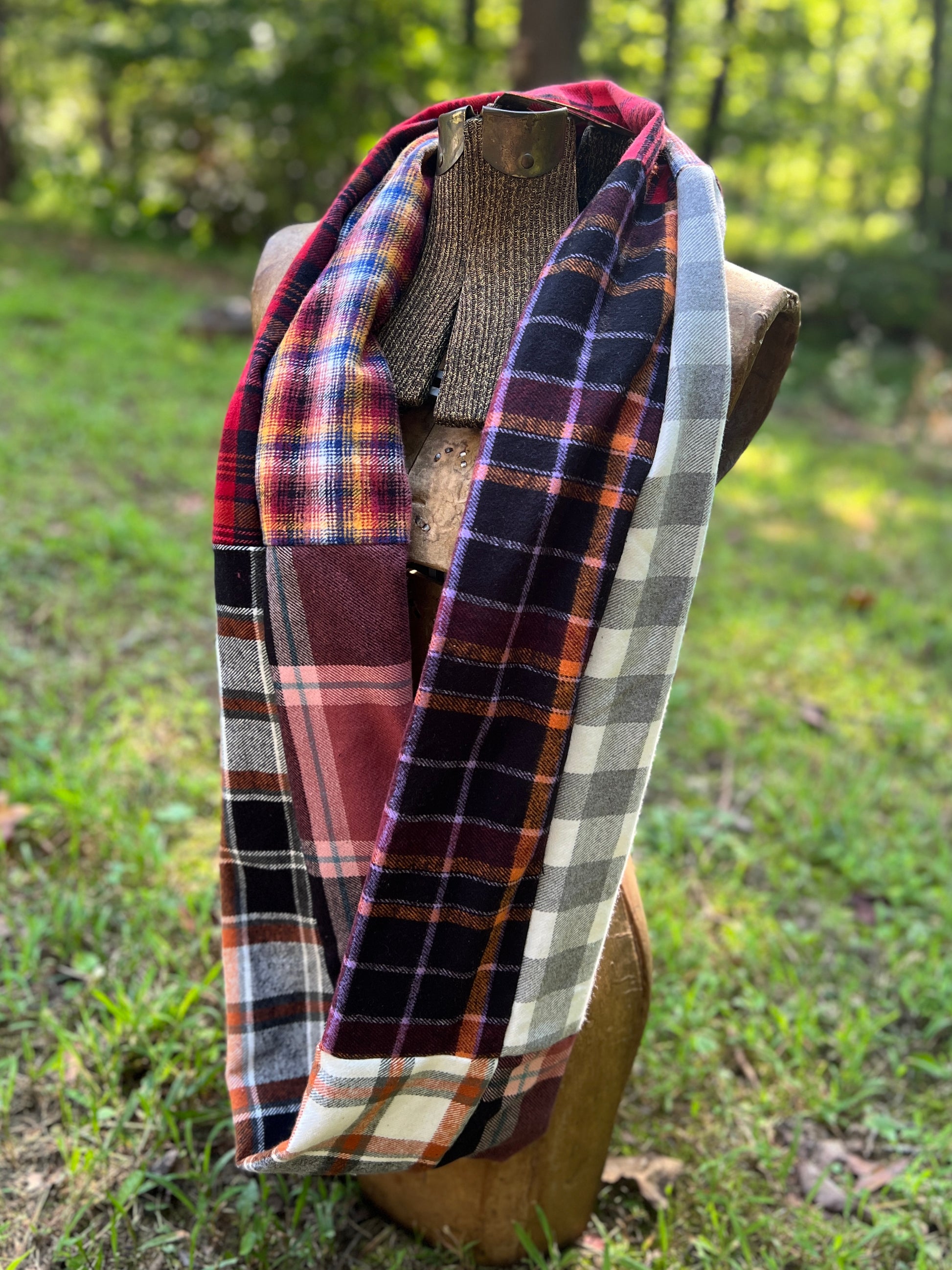 A multicolor patchwork plaid scarf draped on a wooden mannequin torso outdoors on a grassy lawn with trees in the background.