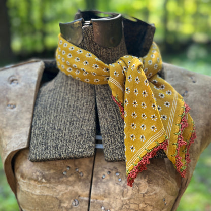 A mustard yellow scarf with a white and green daisy pattern and red floral fringe, styled on a medieval-style wooden armor display against a blurred green outdoor background.