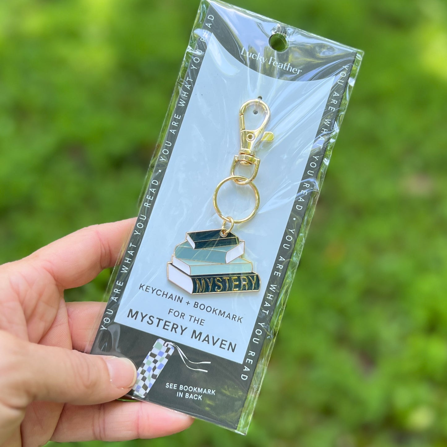 A hand holding a packaged keychain and bookmark set. The product is an enamel book charm with gold-tone hardware, attached to a light blue backing card that reads 'Lucky Feather' and 'Keychain + Bookmark for the Mystery Maven.