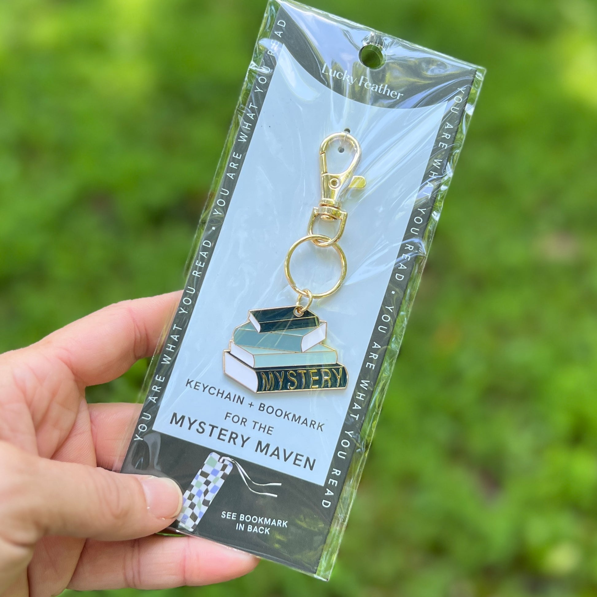 A hand holding a packaged keychain and bookmark set. The product is an enamel book charm with gold-tone hardware, attached to a light blue backing card that reads 'Lucky Feather' and 'Keychain + Bookmark for the Mystery Maven.