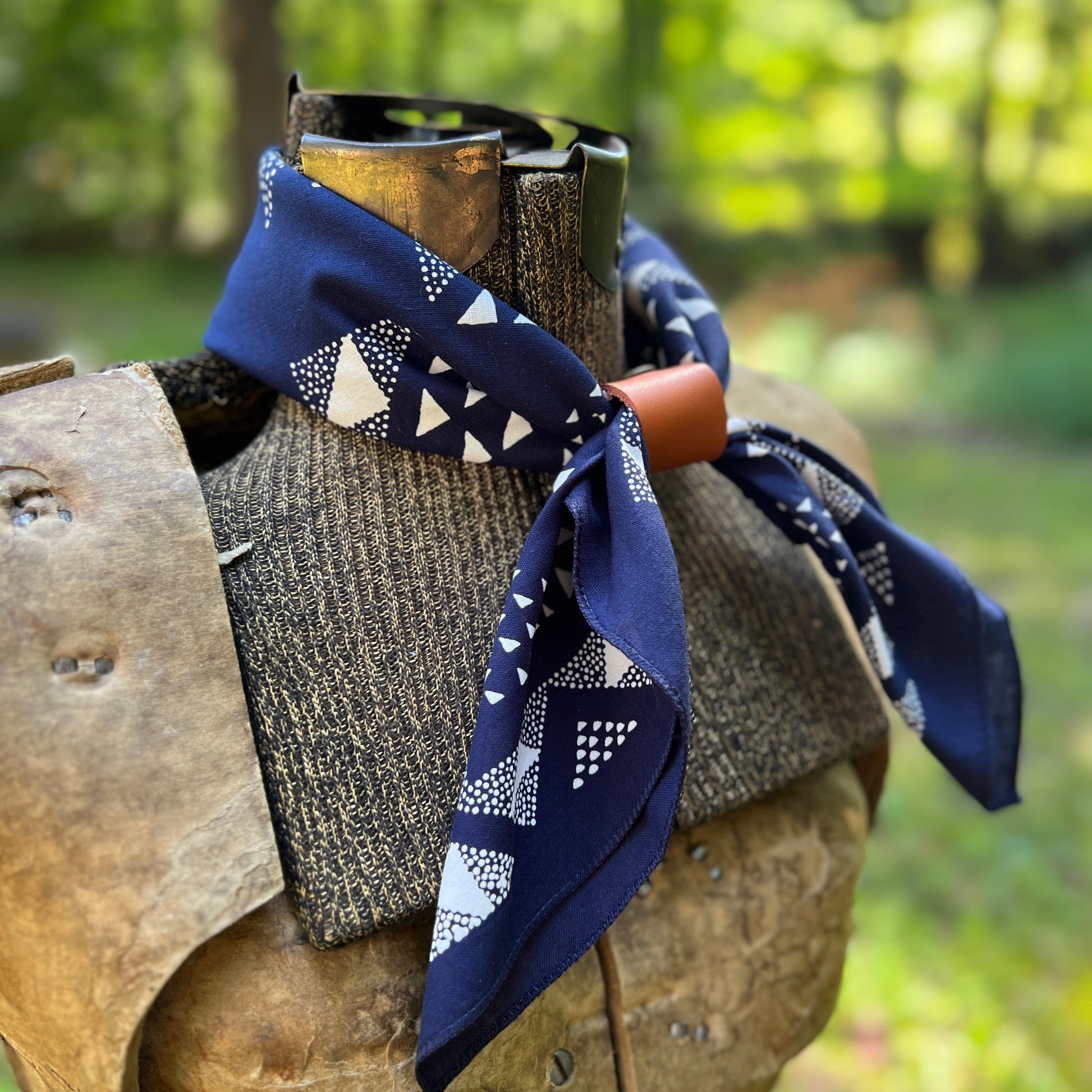 A navy blue bandana with a white geometric pattern is tied around the neck of a rustic wooden mannequin torso, set against a blurred green forest background.