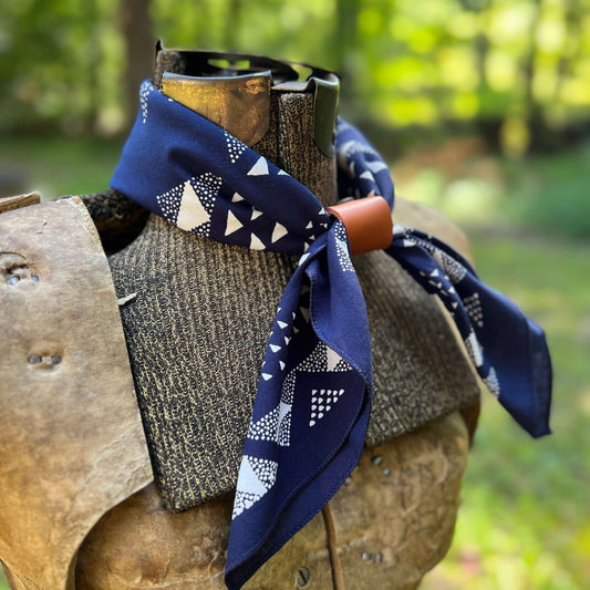 A navy blue bandana with a white geometric pattern is tied around the neck of a rustic wooden mannequin torso, set against a blurred green forest background.