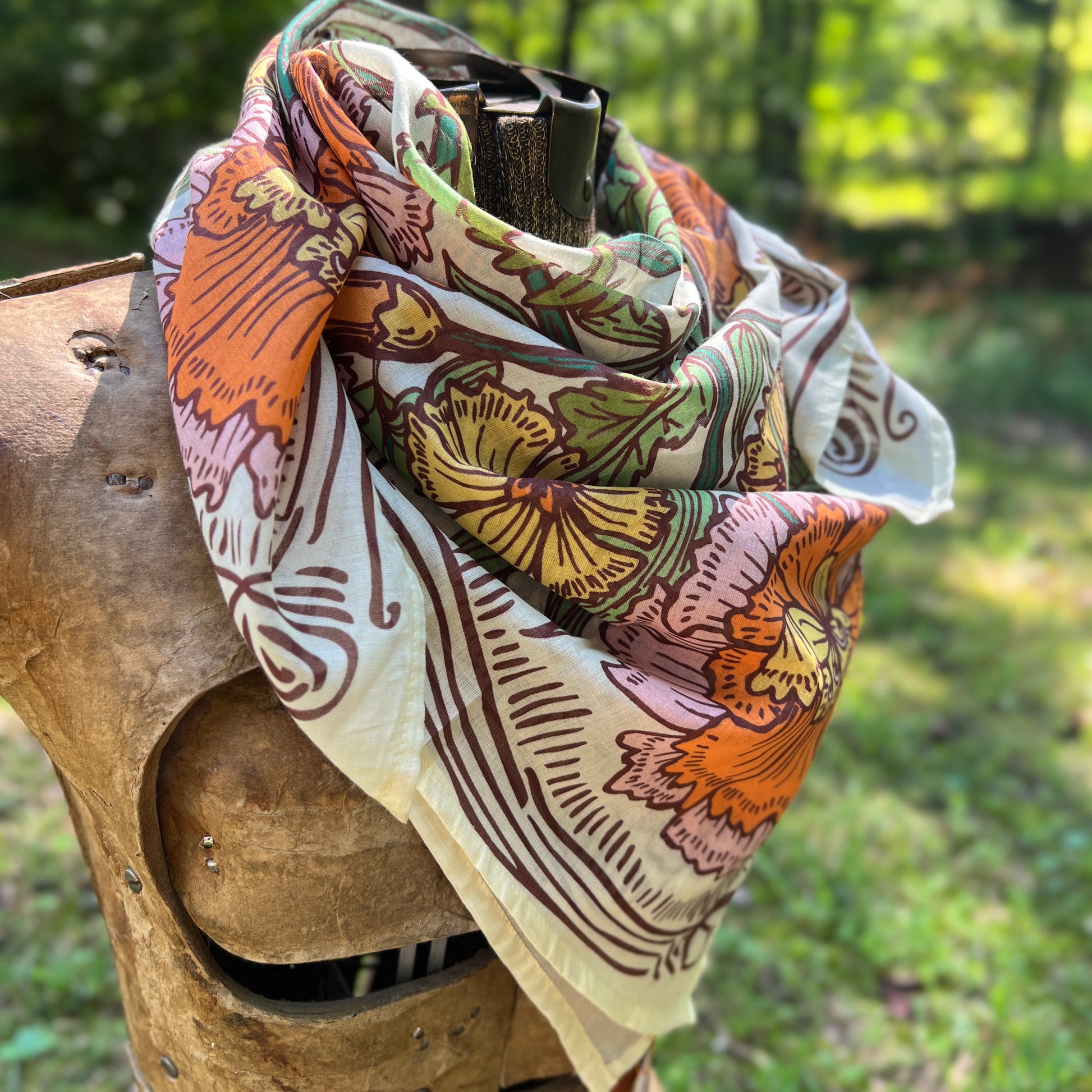 A multicolor floral scarf with orange, yellow, green, and pink patterns on a cream background, draped over a rustic wooden mannequin outdoors.