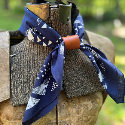 A navy blue patterned neck scarf with white geometric shapes, tied around a rustic brown wooden collar on an outdoor mannequin bust with a blurred green background.