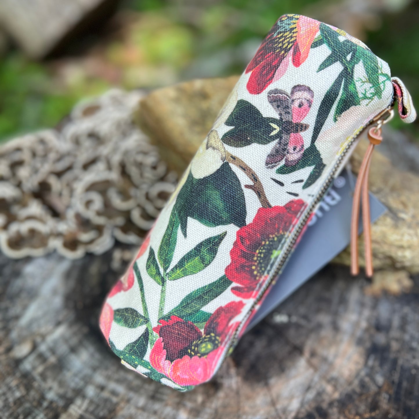 A fabric zippered pencil case with a colorful floral and butterfly print, resting on a rustic wooden surface with a lace doily in the background.
