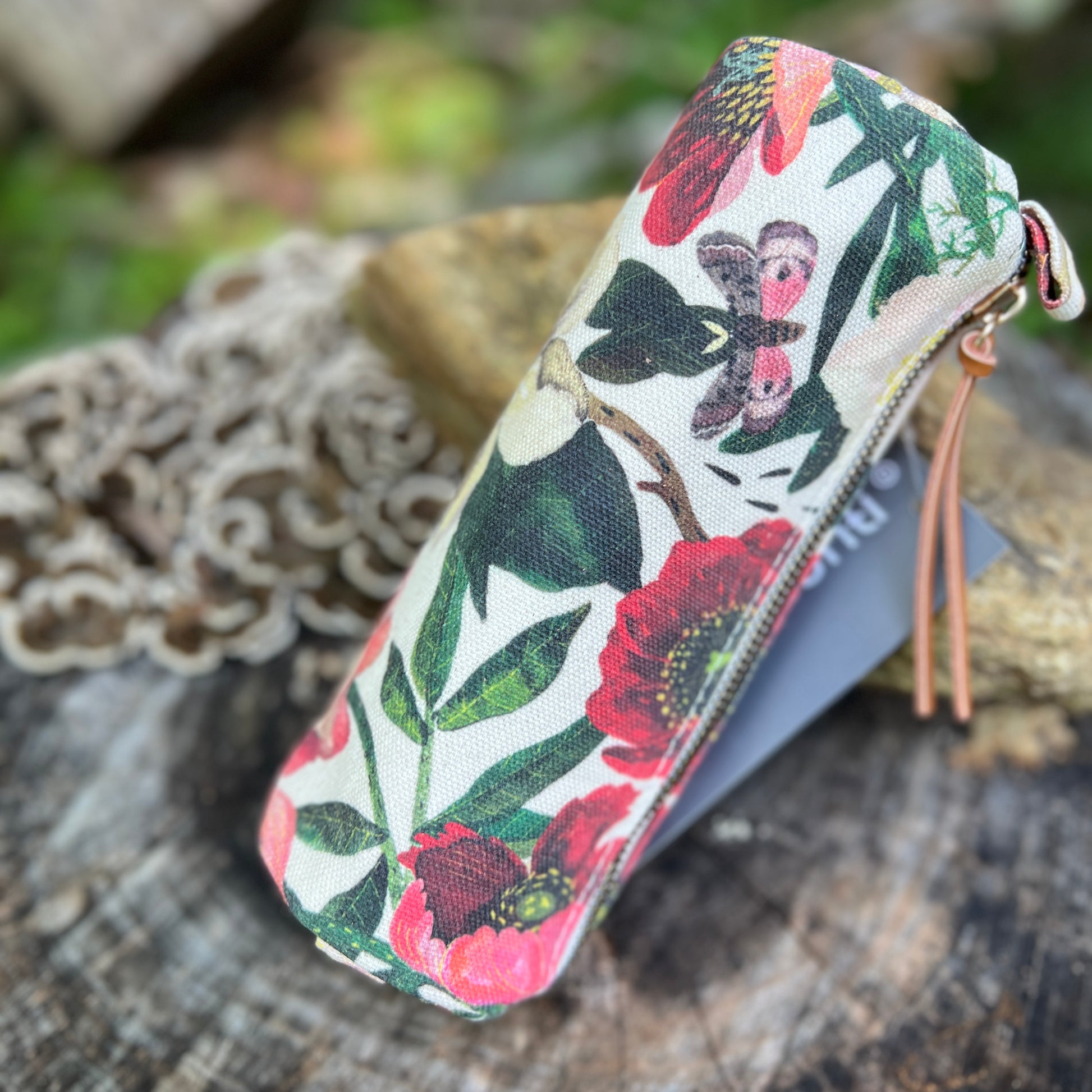 A fabric zippered pencil case with a colorful floral and butterfly print, resting on a rustic wooden surface with a lace doily in the background.