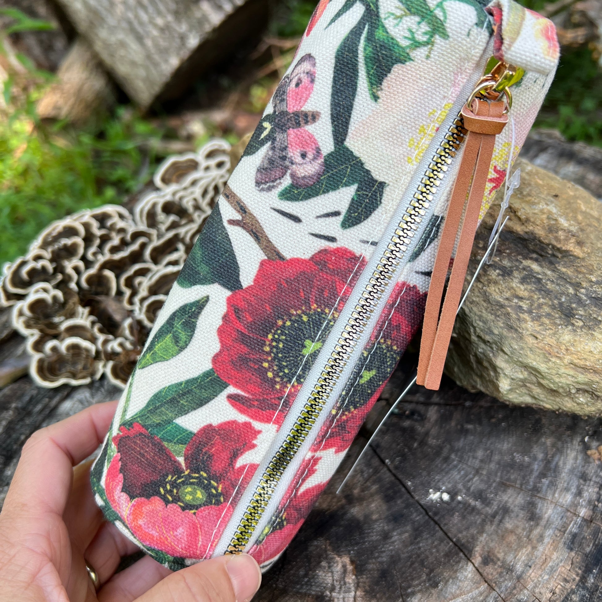 A hand holds a rectangular canvas pouch with a floral print featuring red poppies, green leaves, and a butterfly. The pouch has a gold zipper, a tan leather wrist strap, and is photographed outdoors on a rustic wooden surface with mushrooms and greenery in the background.
