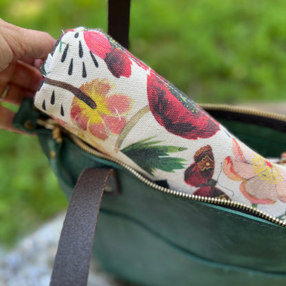 A close-up of a hand holding a rolled fabric pouch with a colorful floral print inside an open dark green bag with a gold zipper.