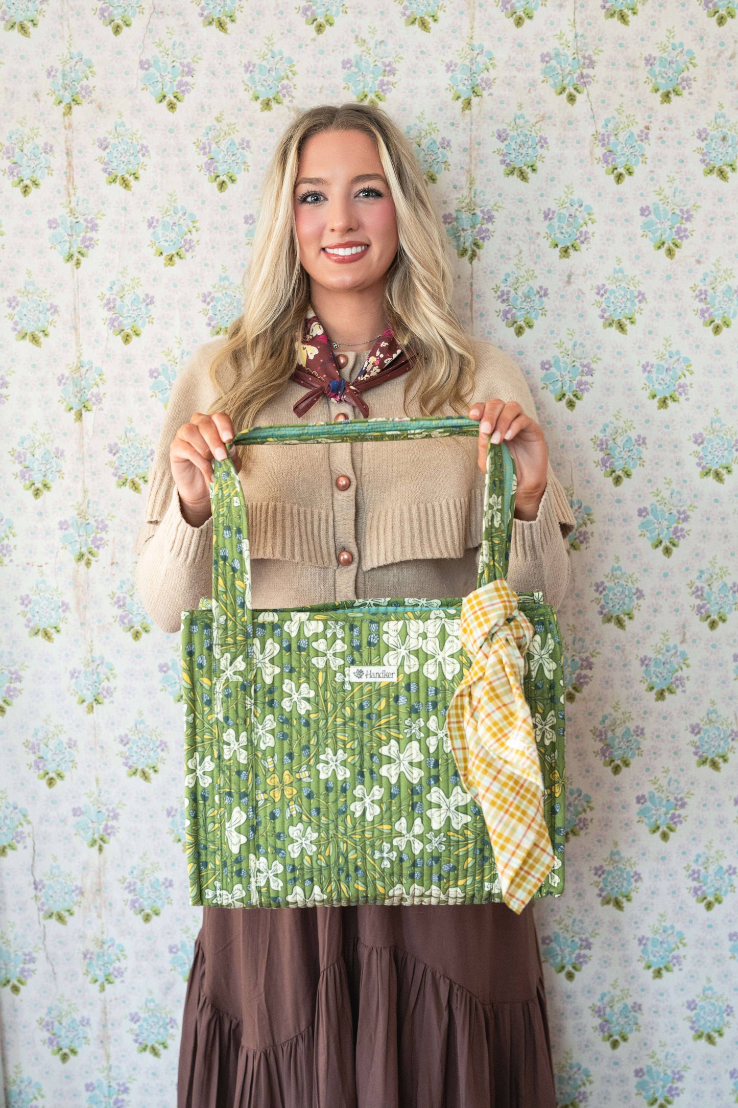 Woman holding a green floral-patterned bag against a floral wallpaper background