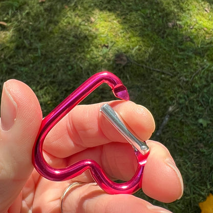 A close-up of a hand holding a small metallic heart-shaped carabiner outdoors on a grassy background. The carabiner has a bright pink body with a silver metal latch.