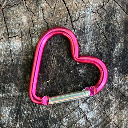 A bright pink heart-shaped metal carabiner with a silver locking mechanism, placed on a textured dark wood surface.