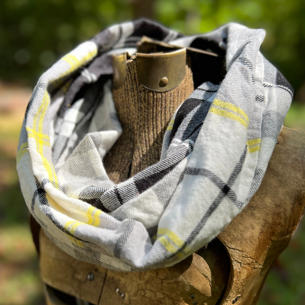 A plaid infinity scarf in white, black, and yellow, draped around a rustic wooden display stand outdoors with a blurred green forest background.