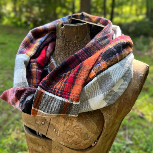 A plaid scarf in red, orange, black, white, and gray draped on a rustic wooden display stand outdoors with a blurred green forest background.