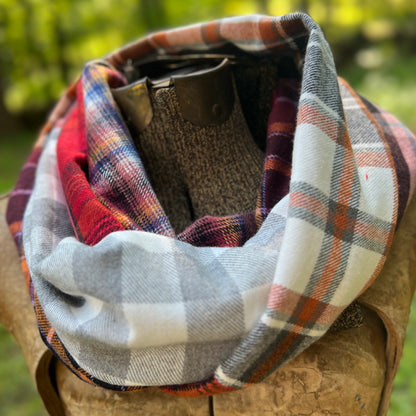 A plaid infinity scarf in a mix of gray, white, red, and blue tones, draped around a brown leather boot on a rustic wooden stand with a blurred green outdoor background.