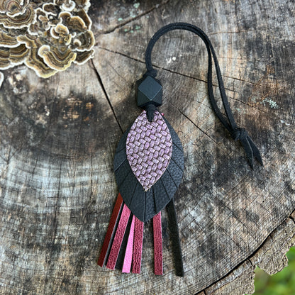 A handmade leather tassel ornament featuring a textured pink and black leaf-shaped pendant with hanging leather strips in pink, burgundy, and black, displayed on a rustic wood log background.