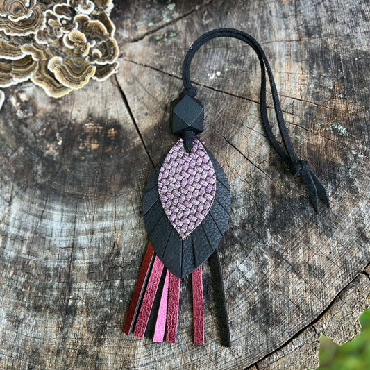 A handmade leather tassel ornament featuring a textured pink and black leaf-shaped pendant with hanging leather strips in pink, burgundy, and black, displayed on a rustic wood log background.