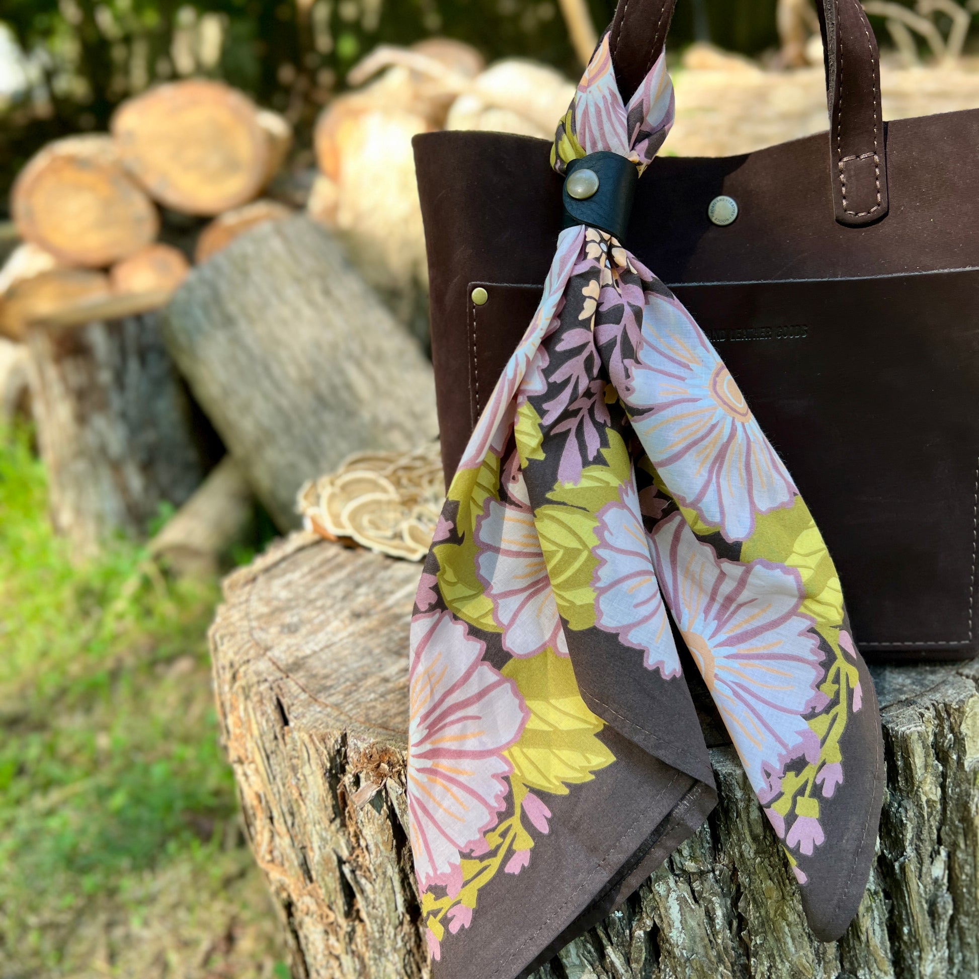 A dark brown leather tote bag with a floral cotton scarf tied around its handle, set outdoors on a rustic tree stump with blurred logs and grass in the background.