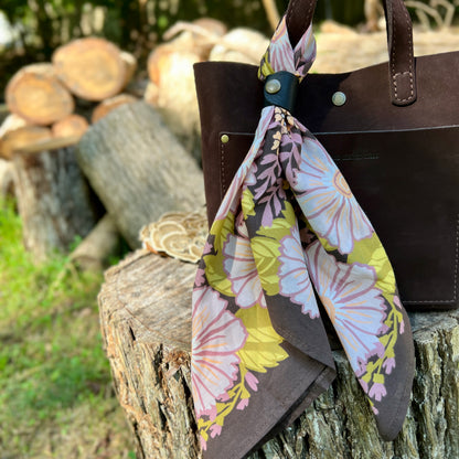 A dark brown leather tote bag with a floral cotton scarf tied around its handle, set outdoors on a rustic tree stump with blurred logs and grass in the background.