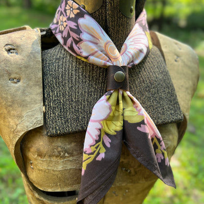 A floral cotton neck scarf with pink, yellow, and white flowers on a dark purple background, styled around a medieval-style wooden torso display outdoors.