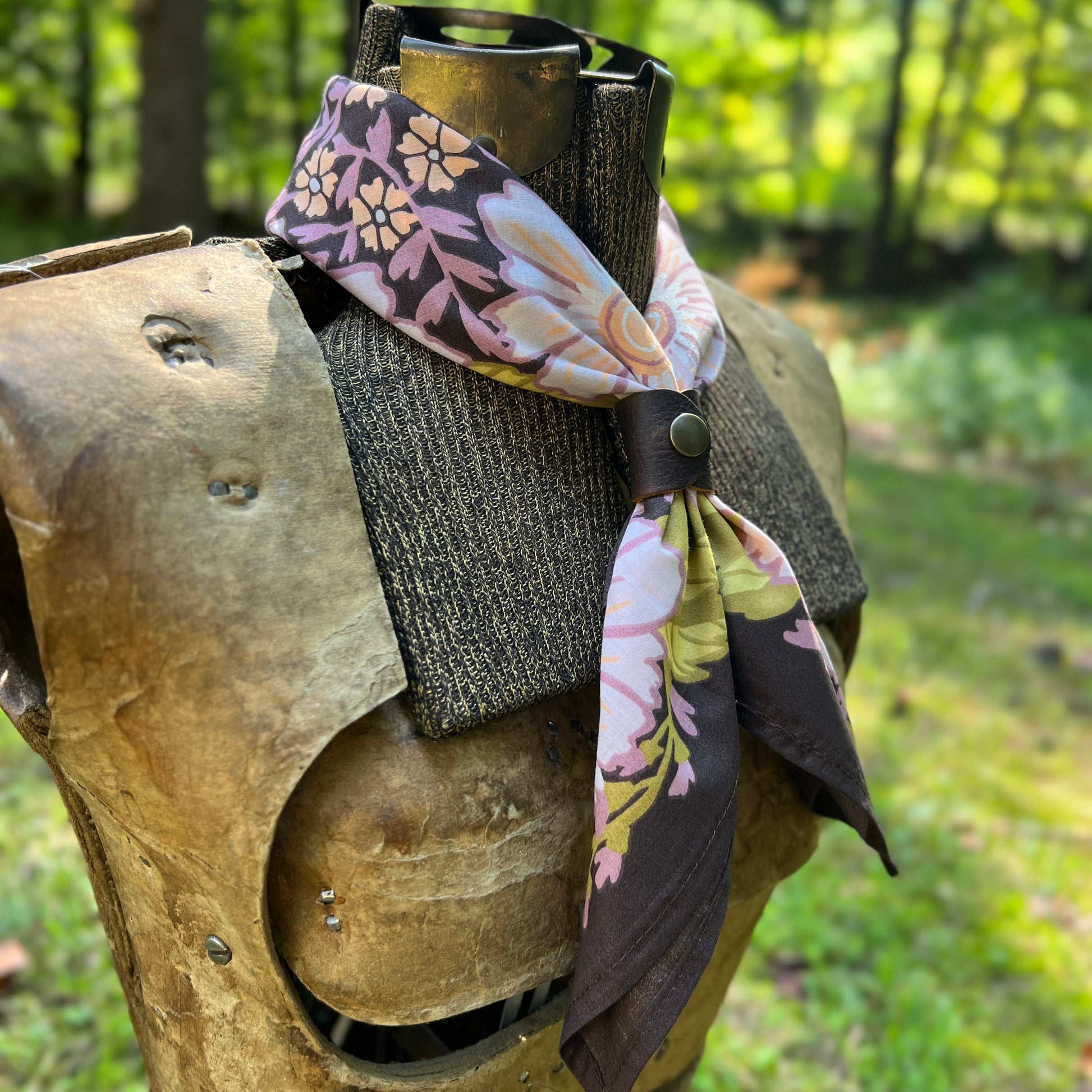 A floral neck scarf with a dark brown base and pink, yellow, and white botanical patterns, styled on a medieval-style wooden armor display outdoors in a grassy wooded setting.