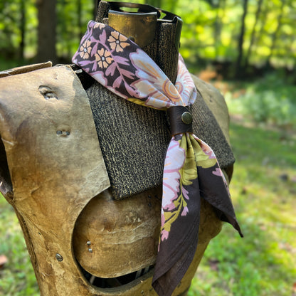 A floral neck scarf with a dark brown base and pink, yellow, and white botanical patterns, styled on a medieval-style wooden armor display outdoors in a grassy wooded setting.
