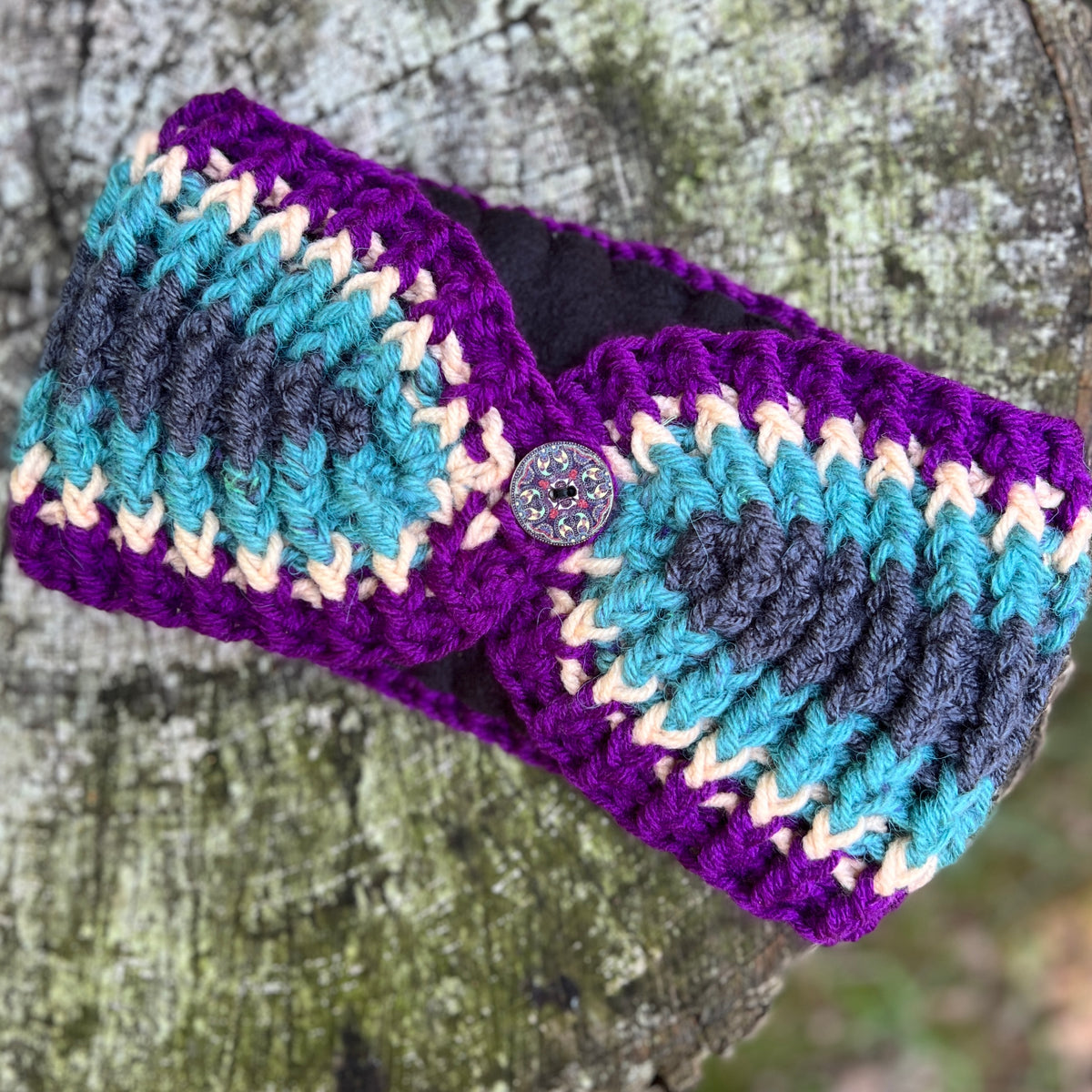 A handmade crocheted headband with a purple base, teal, navy, and cream chevron stripes, featuring a decorative button closure, shown on a textured tree trunk background.