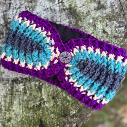 A handmade crocheted headband with a purple base, teal, navy, and cream chevron stripes, featuring a decorative button closure, shown on a textured tree trunk background.