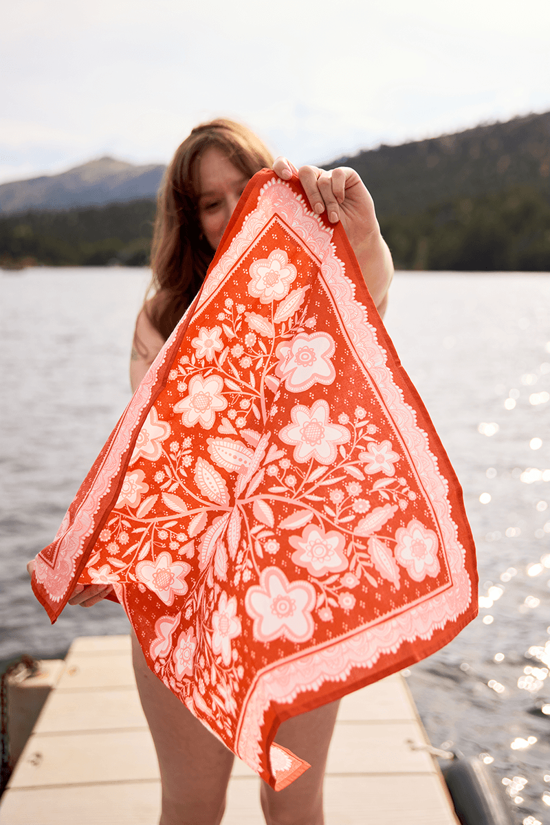 Person holding a red floral scarf by a body of water with mountains in the background