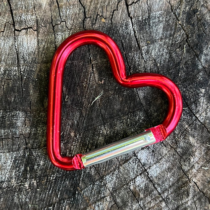 A red heart-shaped carabiner with a silver metal slide, placed on a textured, weathered wood surface.
