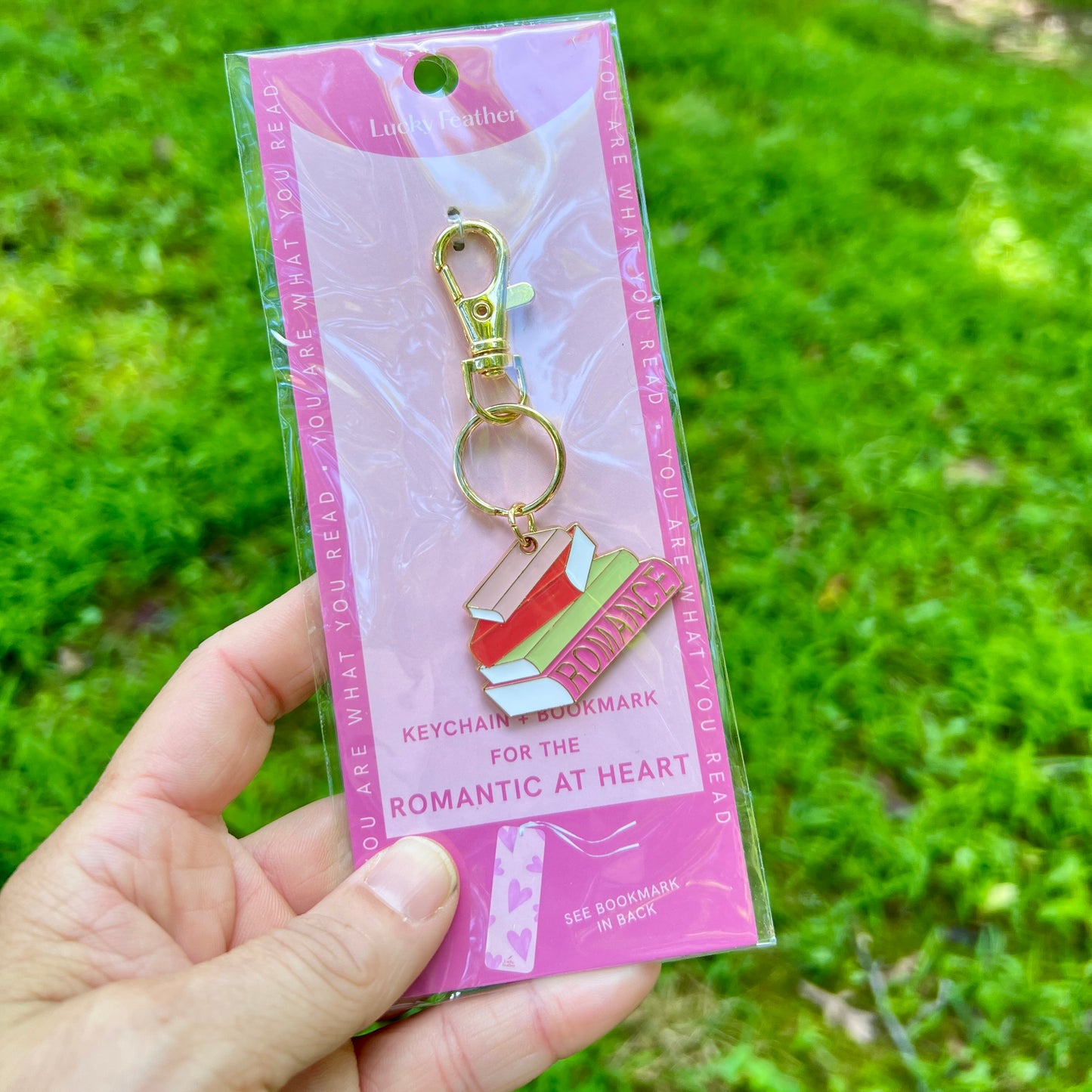 A hand holding a clear plastic blister pack containing a keychain charm shaped like a book with a pink heart and green leaves, set against a blurred green grass background.